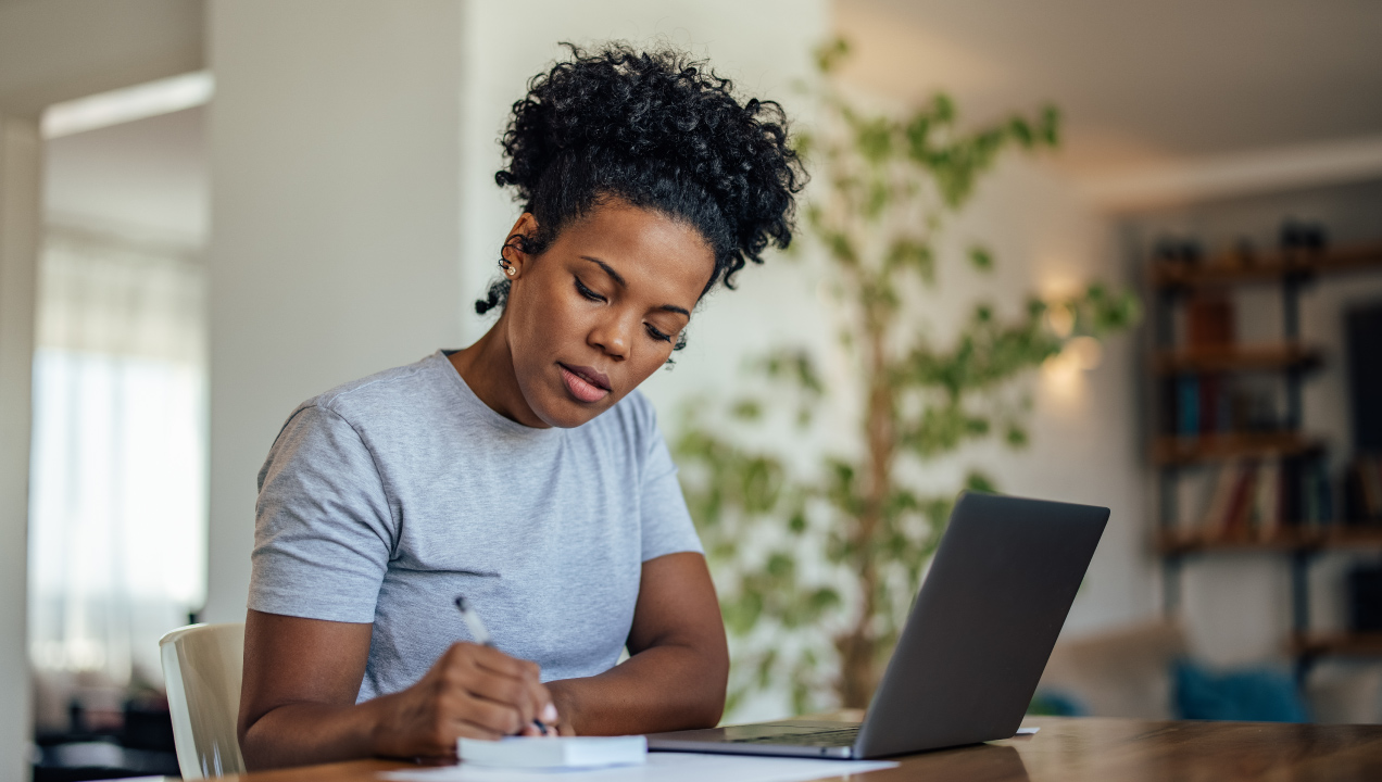 Senhora sentada numa secretária com o seu computador e a escrever num bloco de notas